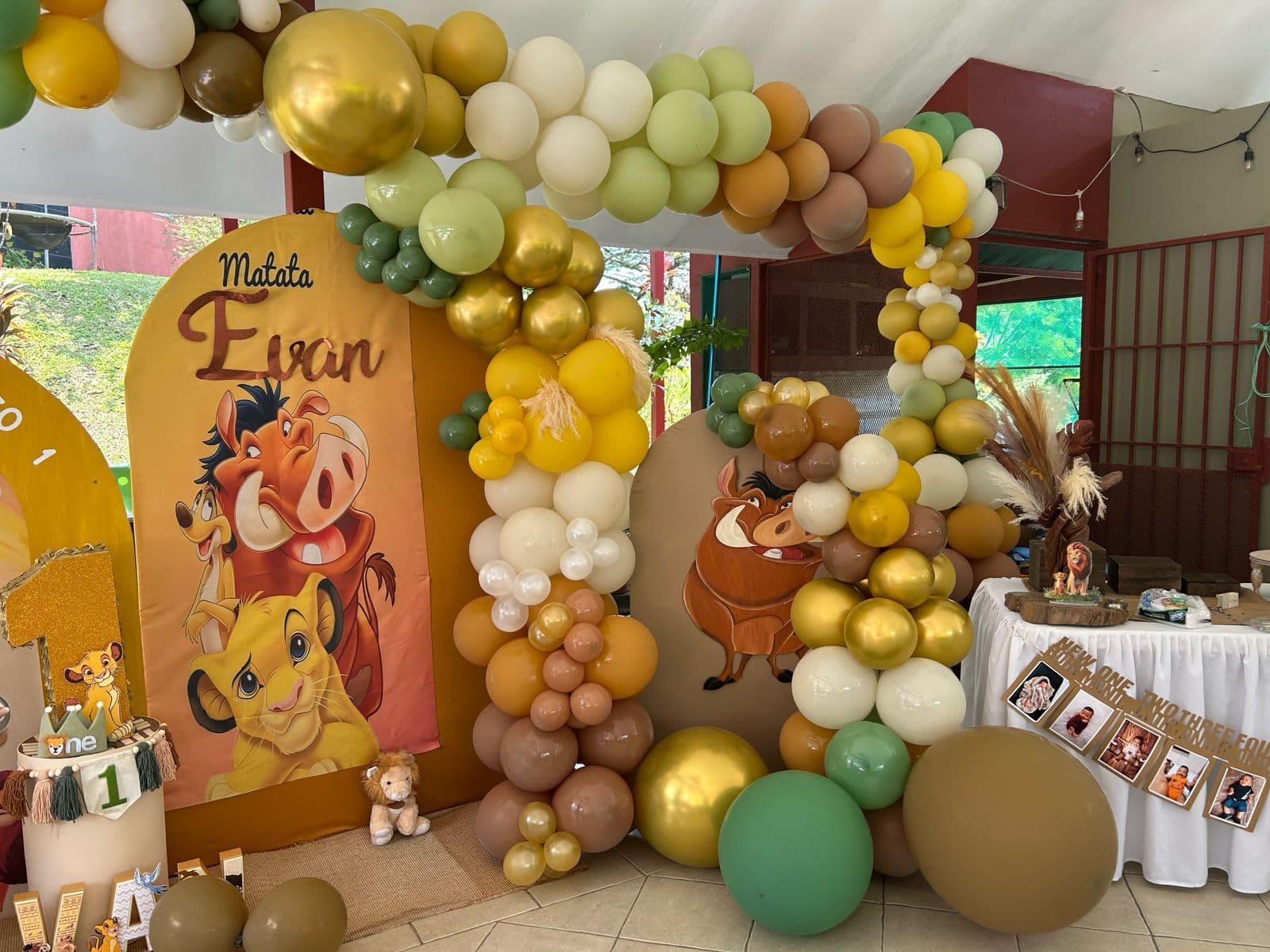 Decoración de primer cumpleaños de El Rey León con paneles de Simba, Timón y Pumba y un arco de globos verdes, amarillos y dorados