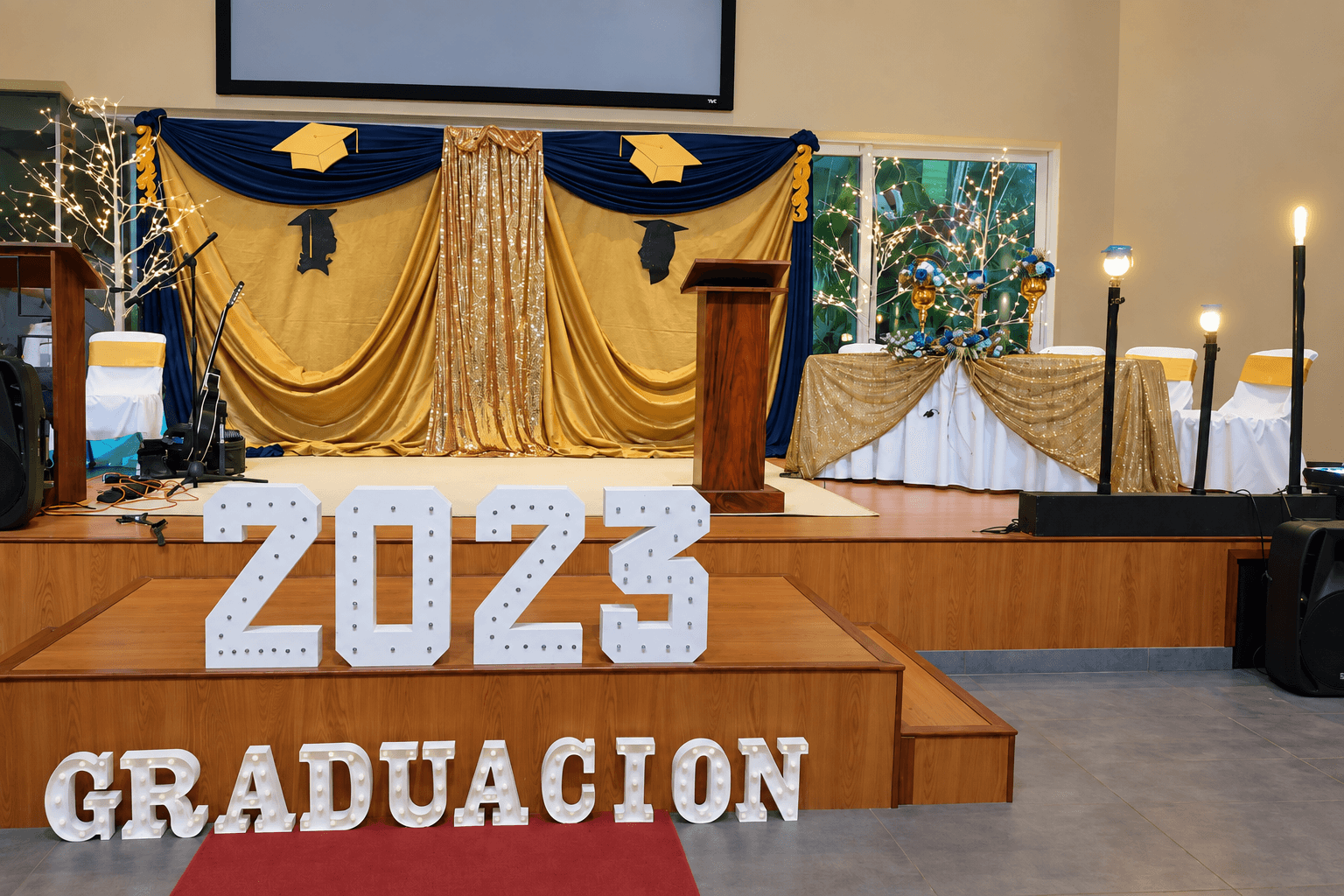 Escenario de graduación con telas azules y doradas, arreglos florales y letras luminosas de graduación 2023